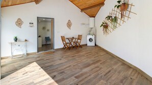 Een kamer met een houten plafond, een wasmachine, een eettafel met stoelen en een kleine tafel met een plant.