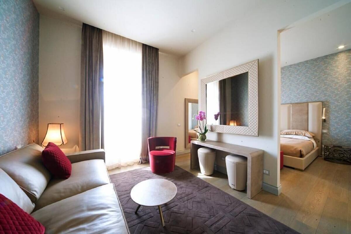 A modern hotel room with a sofa, a small round table, a red chair, a bedside table with a vase of flowers, a large mirror, and a bed.