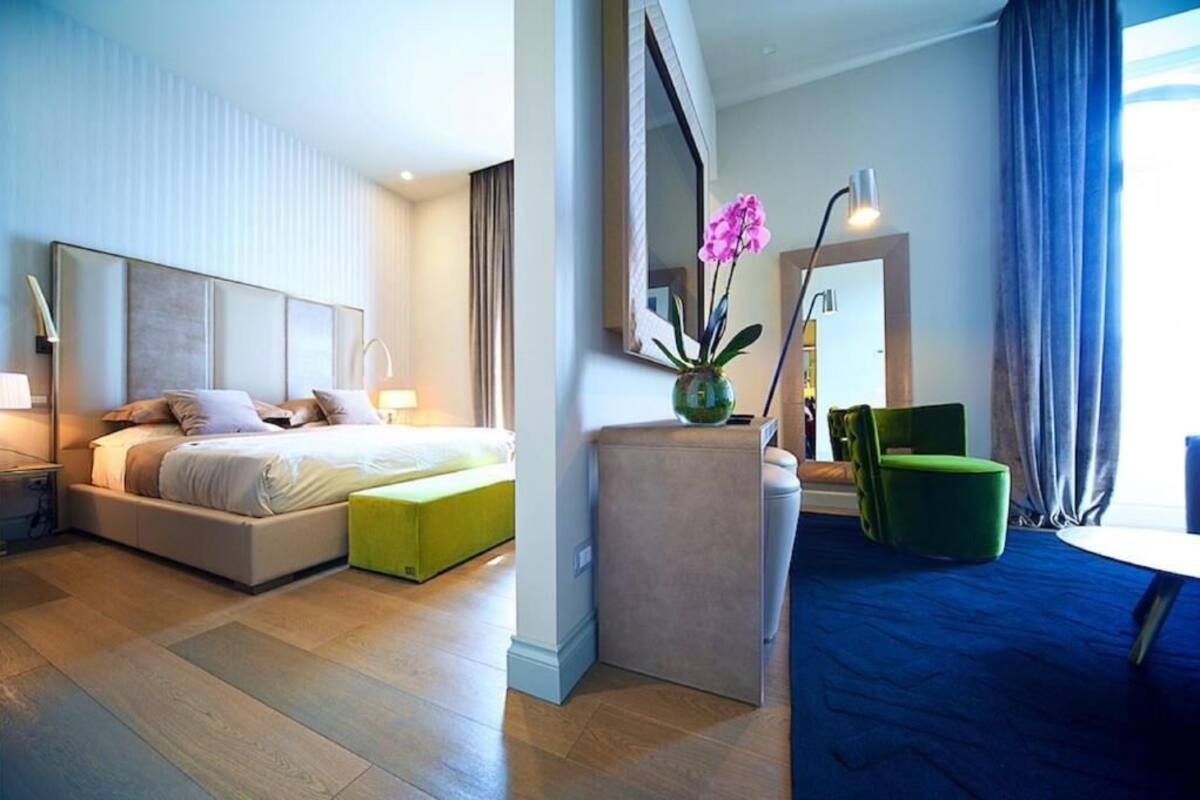 A modern hotel room with a large bed, a green ottoman, a small table, and a mirror.