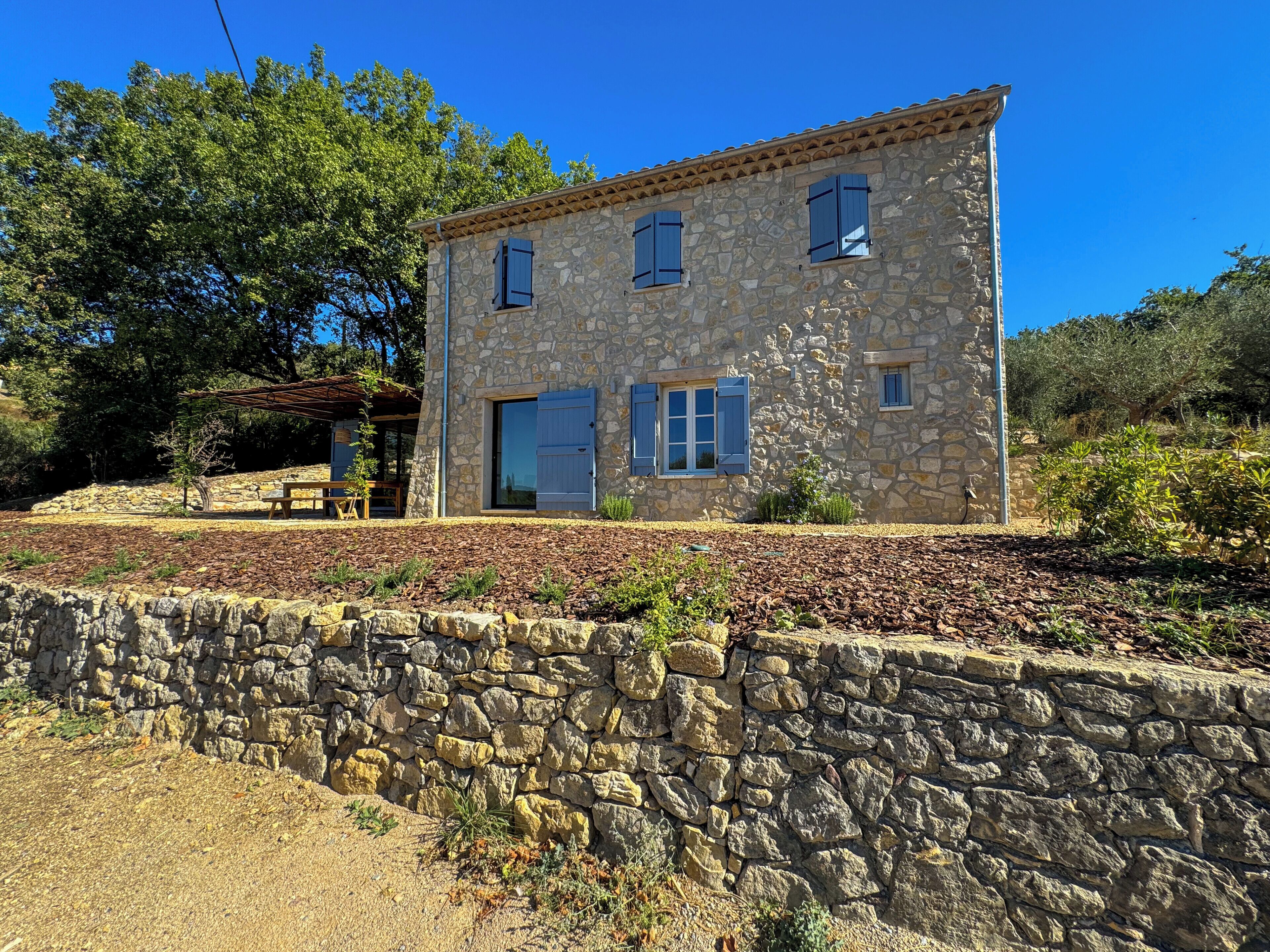 Une maison en pierre avec un muret en pierre devant, un banc et une pergola.