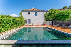 Une piscine avec une terrasse en bois, un bâtiment doté d’un toit en tuiles et une haie.
