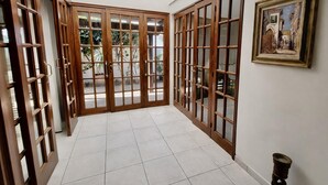 Un pasillo con puertas de madera y suelo de baldosas, que conduce a un balcón con vistas a la vegetación.