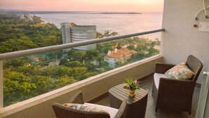 Un balcón con mobiliario de mimbre y una pequeña mesa con flores, con vistas a una ciudad costera con edificios y áreas verdes.
