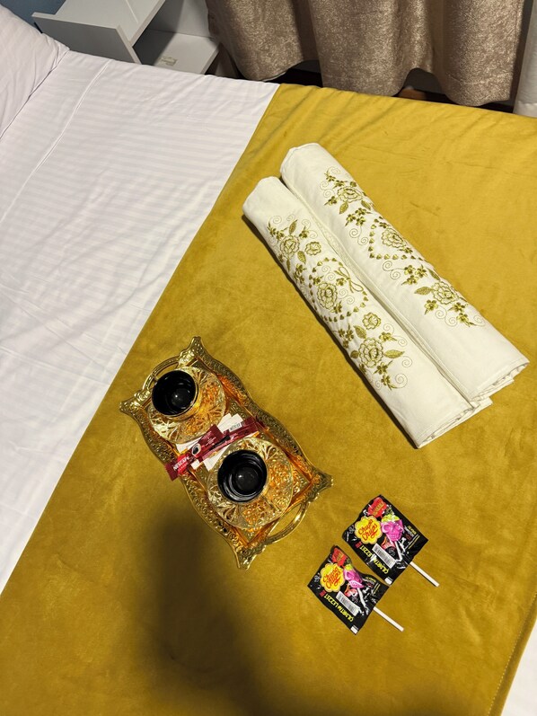 A bed with a white and yellow bedspread, a decorative tray with two black cups and a pink item, and a folded white towel with floral embroidery. - Casafra (Istanbul)
