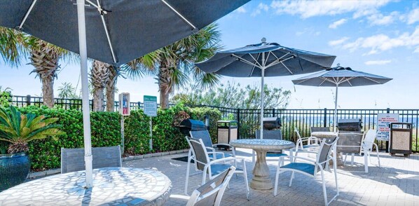 An outdoor patio with round tables and chairs, umbrellas, and a grill area. - Palacio 903 (Pensacola)