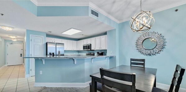 A modern kitchen with white cabinets, a central island, and a dining area with a table and chairs. - Palacio 903 (Pensacola)