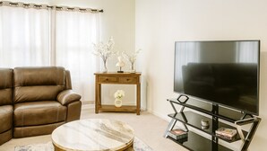 Een woonkamer met een bruine leren bank, een ronde marmeren salontafel, een klein houten bijzettafeltje met een bloevas, en een zwart tv-meubel met een flatscreen televisie.