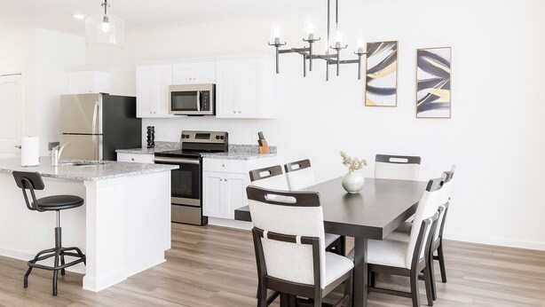Een moderne keuken met witte kastjes, roestvrijstalen apparatuur, een centraal kookeiland met barkrukken, en een eethoek met een donker houten tafel en stoelen.