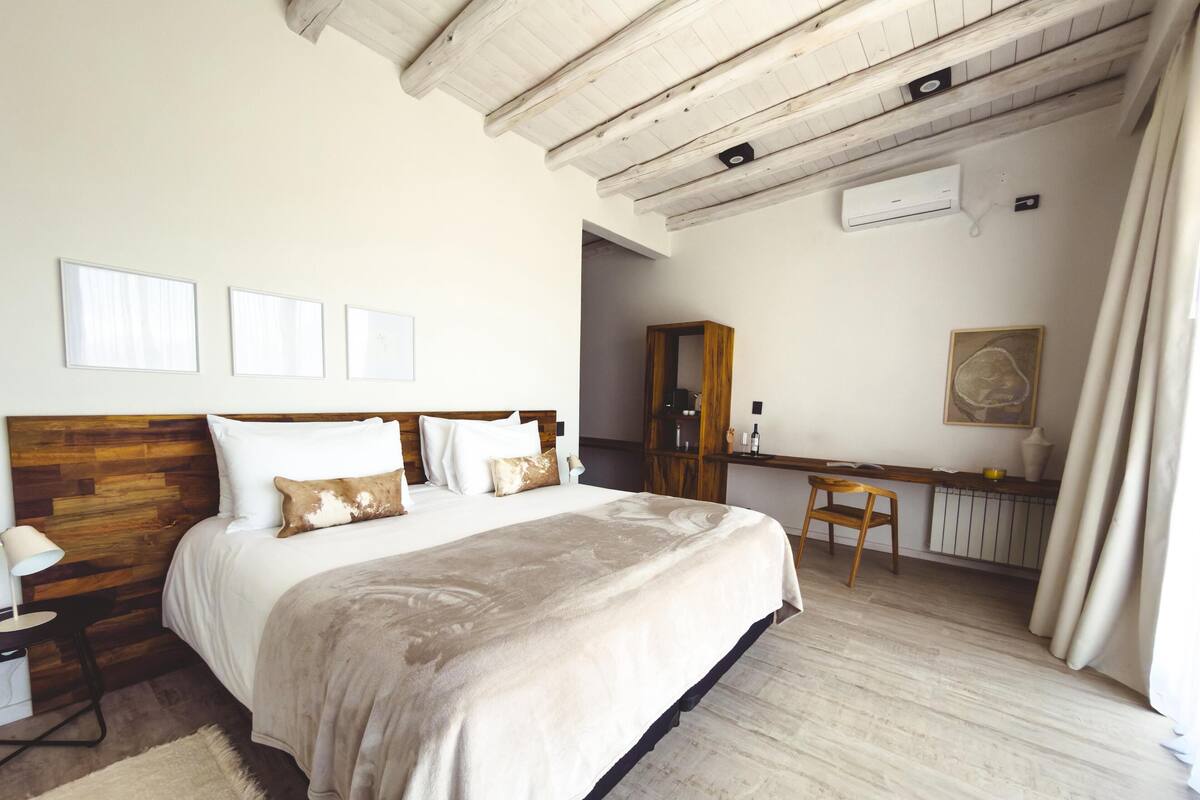 A bedroom with a wooden headboard, a large bed with multiple pillows, a bedside table with a lamp, a wooden desk with a chair, and a radiator.