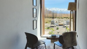 Una habitación con vistas a una cadena montañosa nevada, dos sillas, una mesa con libros y un vaso, y una lámpara.
