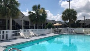 A clear blue swimming pool with white deck chairs and umbrellas, surrounded by palm trees and a white fence. - Best Inn of Manning (Manning)
