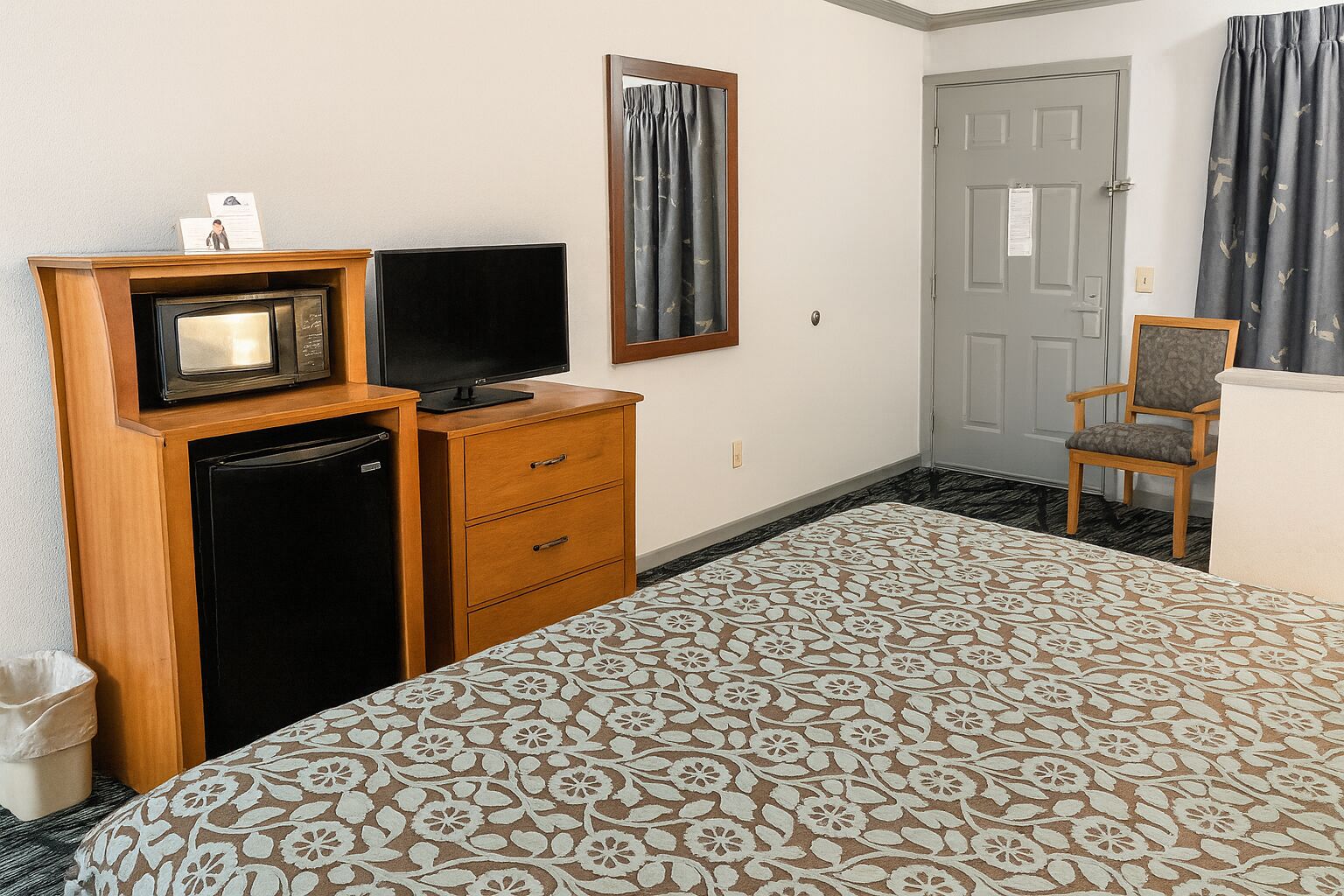 Una habitación de hotel con una cama, televisión, refrigerador, cómoda de madera y una silla.