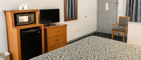 Una habitación de hotel con una cama, televisión, refrigerador, cómoda de madera y una silla.