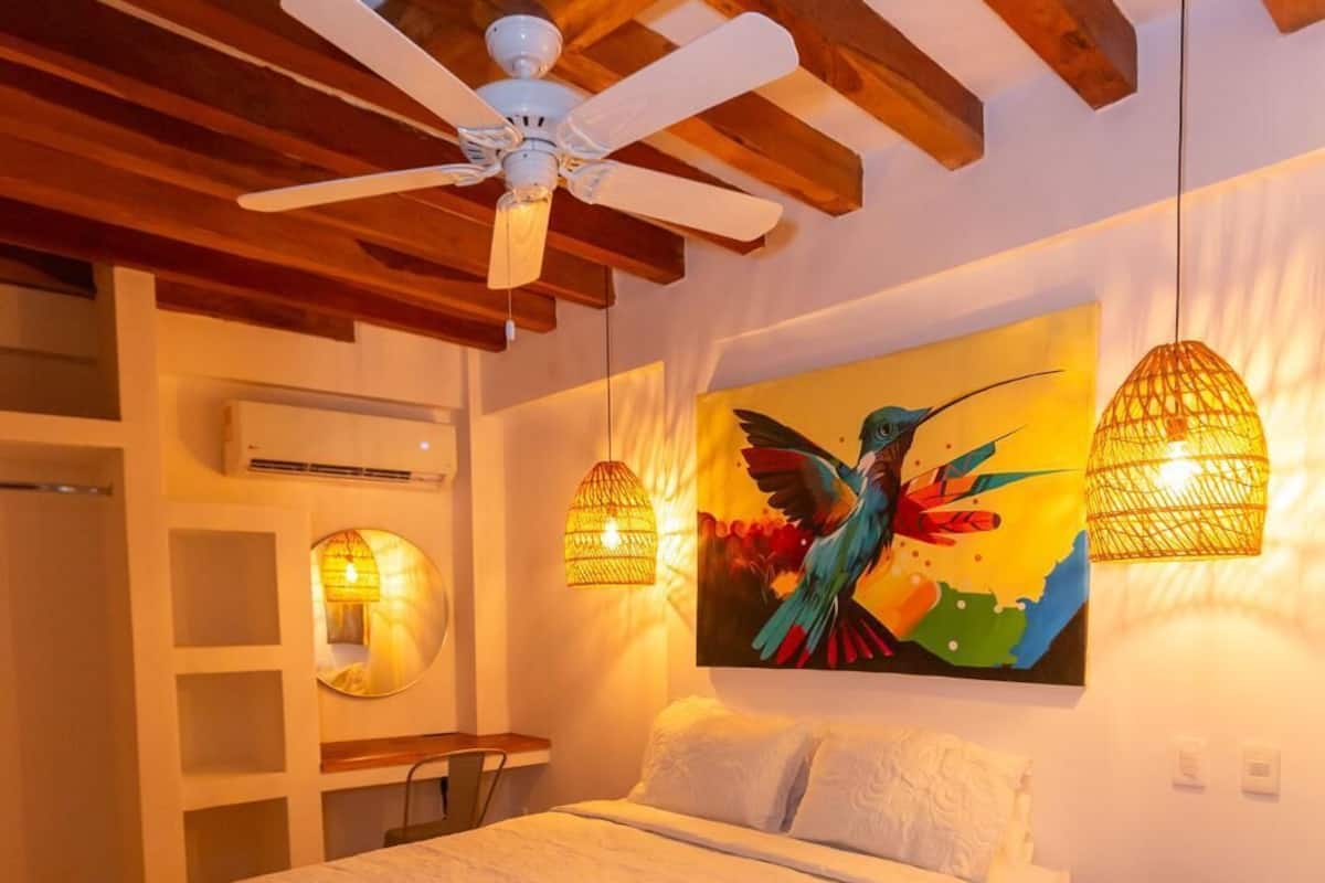A bedroom with a bed, a ceiling fan, a painting, and two wall-mounted lamps.