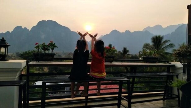 Deux personnes sont assises sur un balcon, d’où l’on peut admirer des montagnes et le soleil couchant à l’horizon.