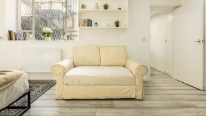A modern living room with a beige sofa, a white radiator, a bookshelf with books and plants, and a view through a window with a metal fence. - Cozy Flat Near Hyde Park! (Greater London)