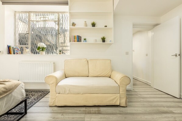A modern living room with a beige sofa, a white radiator, a bookshelf with books and plants, and a view through a window with a metal fence.
