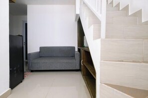 Un pasillo estrecho con un sofá gris, un refrigerador negro y una escalera con escalones color beige.