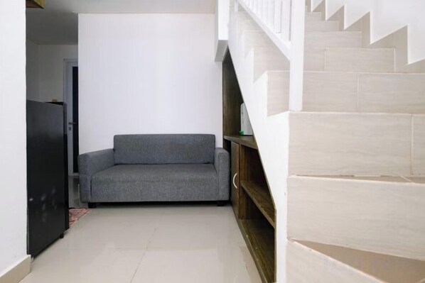 A narrow hallway with a grey sofa, a black refrigerator, and a staircase with beige steps.