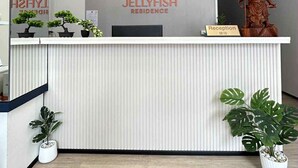 Un mostrador de recepción de diseño moderno, con el logotipo y el nombre 'Jellyfish Residence'. Hay plantas en macetas a cada lado del mostrador, y se ve una estatua de madera a la derecha.