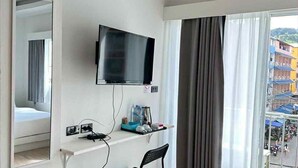 Habitación de hotel con televisión de pantalla plana montada en la pared, kitchenette pequeña con refrigerador, una silla y un cesto de basura.