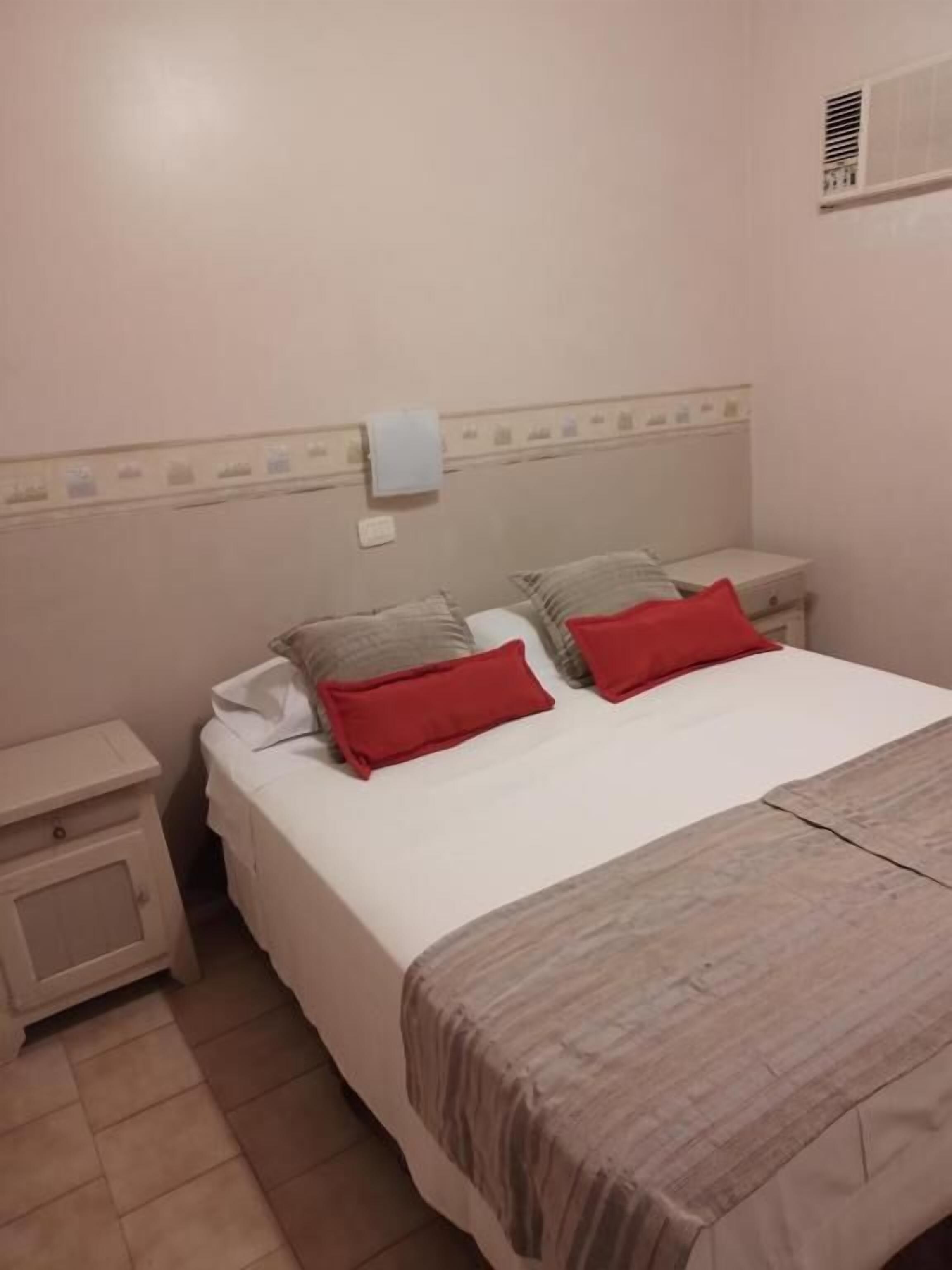 Una cama bien hecha con edredón blanco y dos almohadas rojas, una mesita de noche con lámpara y un respiradero en la pared.