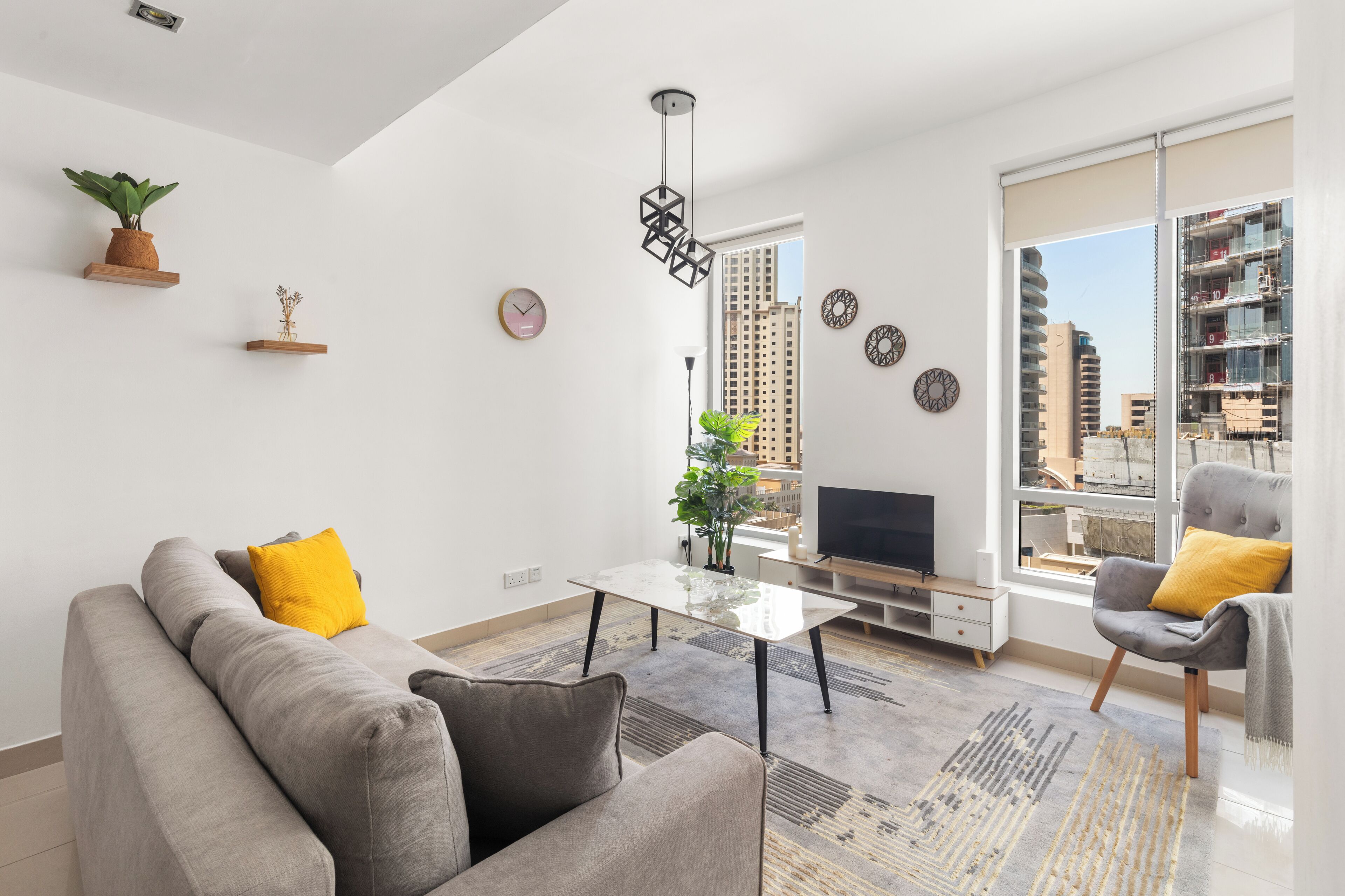 A modern living room with a sofa, armchairs, a coffee table, and a TV. There are plants on shelves and a large window with a city view.