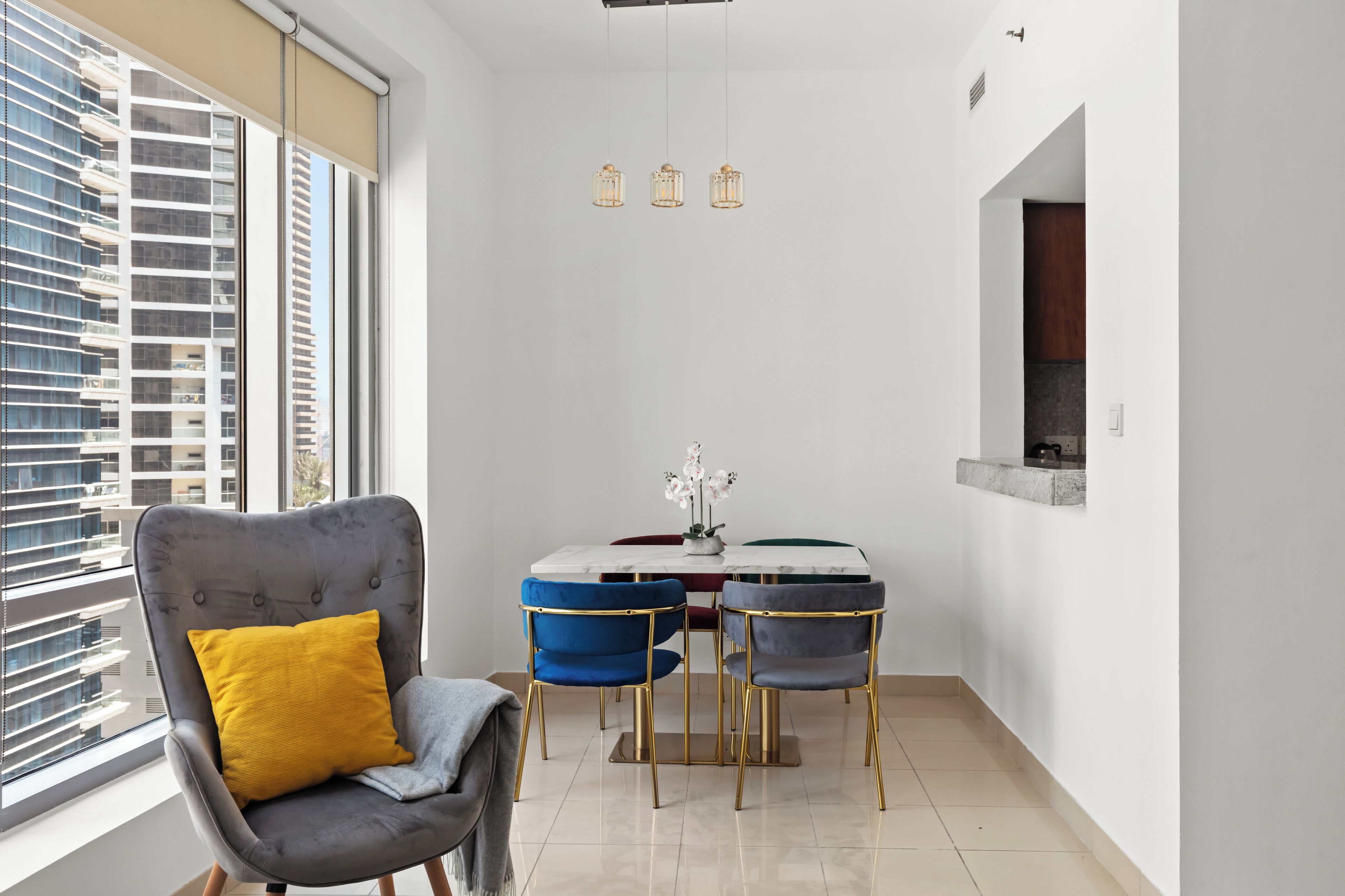 A modern dining area with a marble table and chairs, a grey upholstered chair with a yellow cushion, and a view of a high-rise building through large windows.