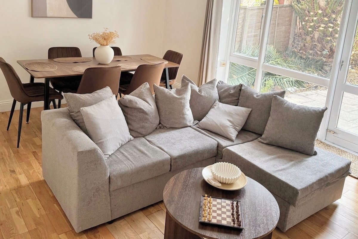 Una sala de estar con un sofá modular, una mesa de comedor con sillas y un tablero de ajedrez sobre una mesita.