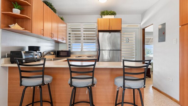 A modern kitchen with a stainless steel refrigerator, a microwave, and a coffee maker on the counter. There are three bar stools and a window with blinds.