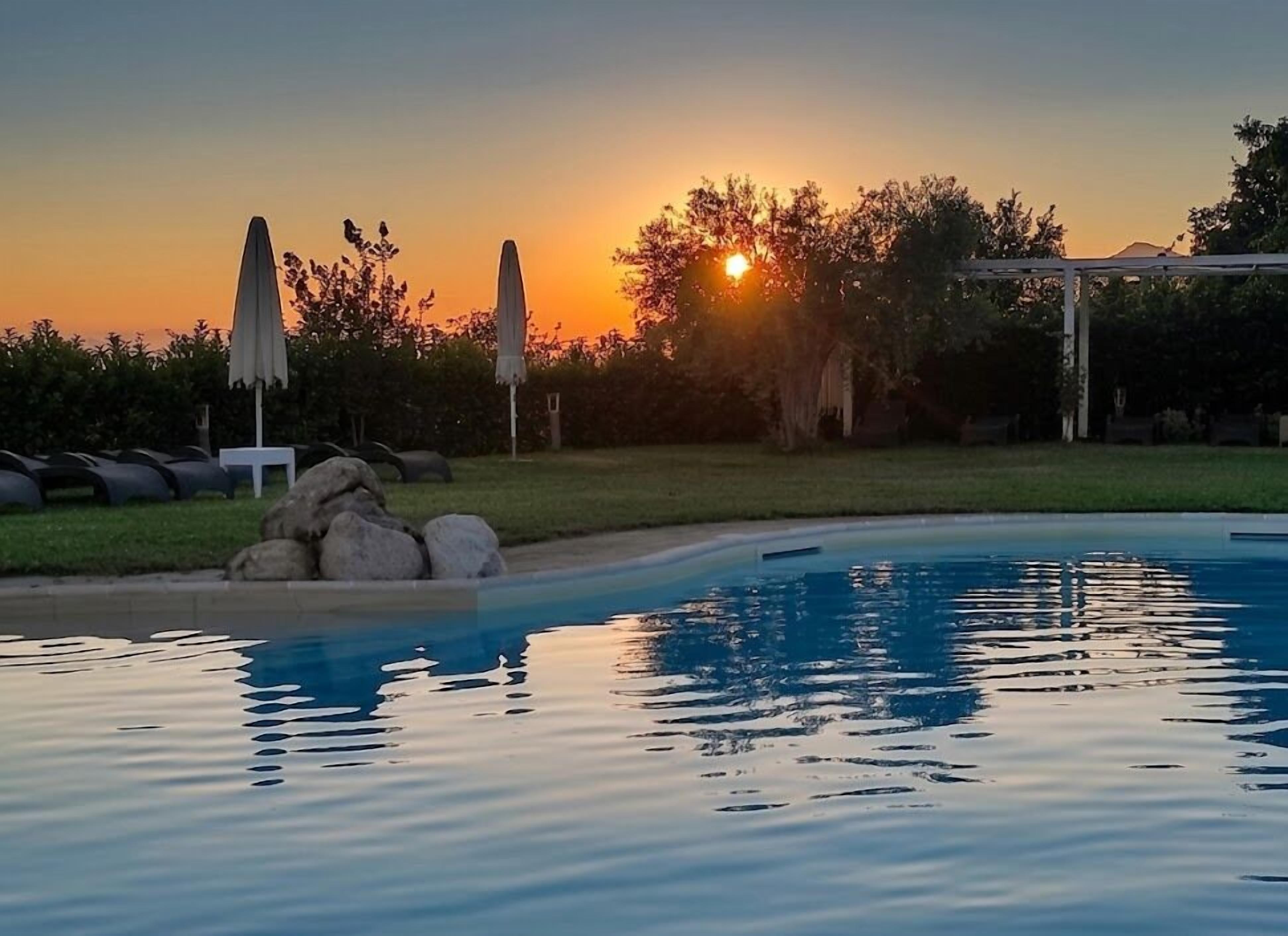 A swimming pool with a sunset in the background, surrounded by rocks and umbrellas.