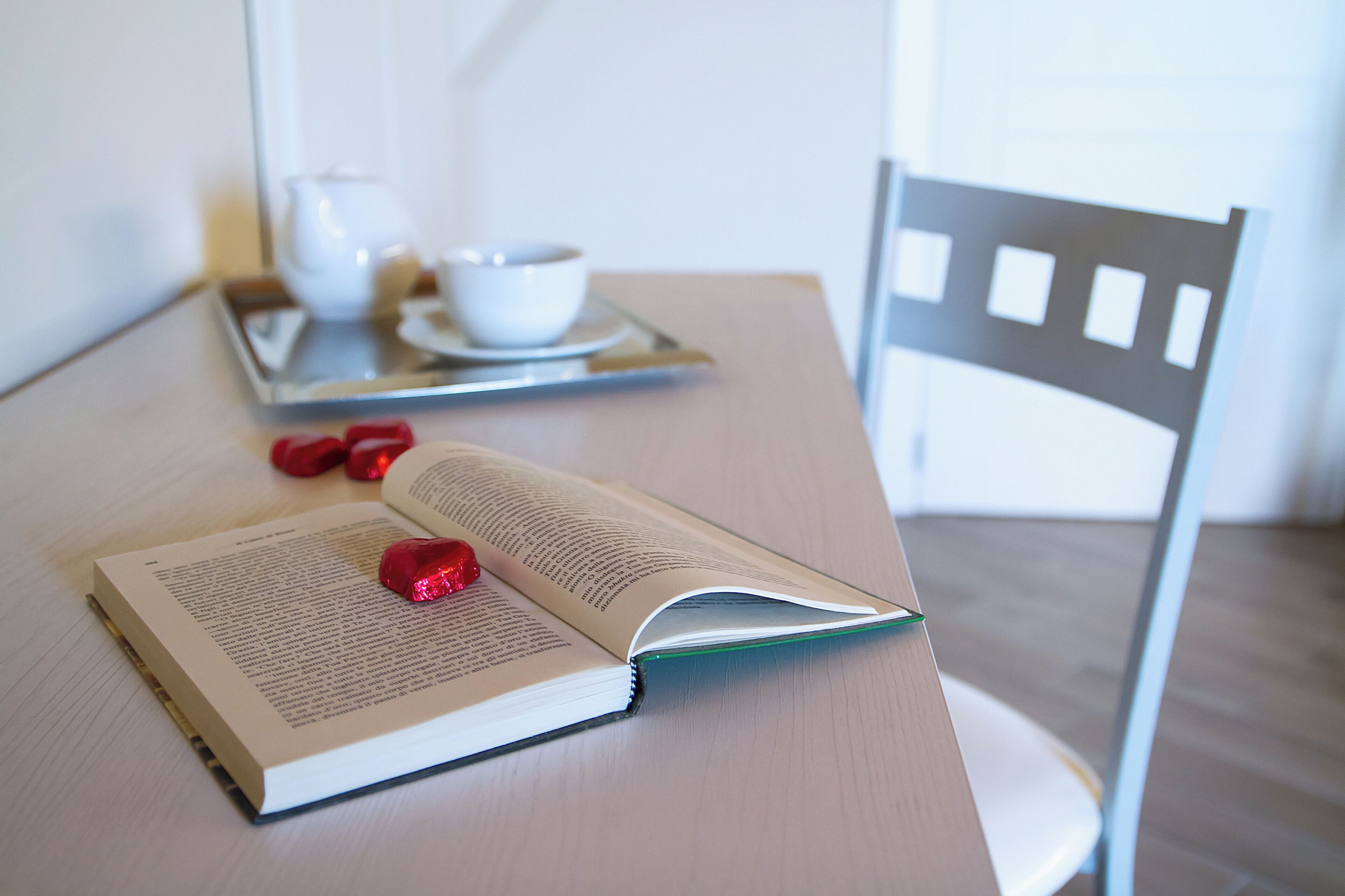 An open book on a table with a tray containing a teapot and cups, and a chair.