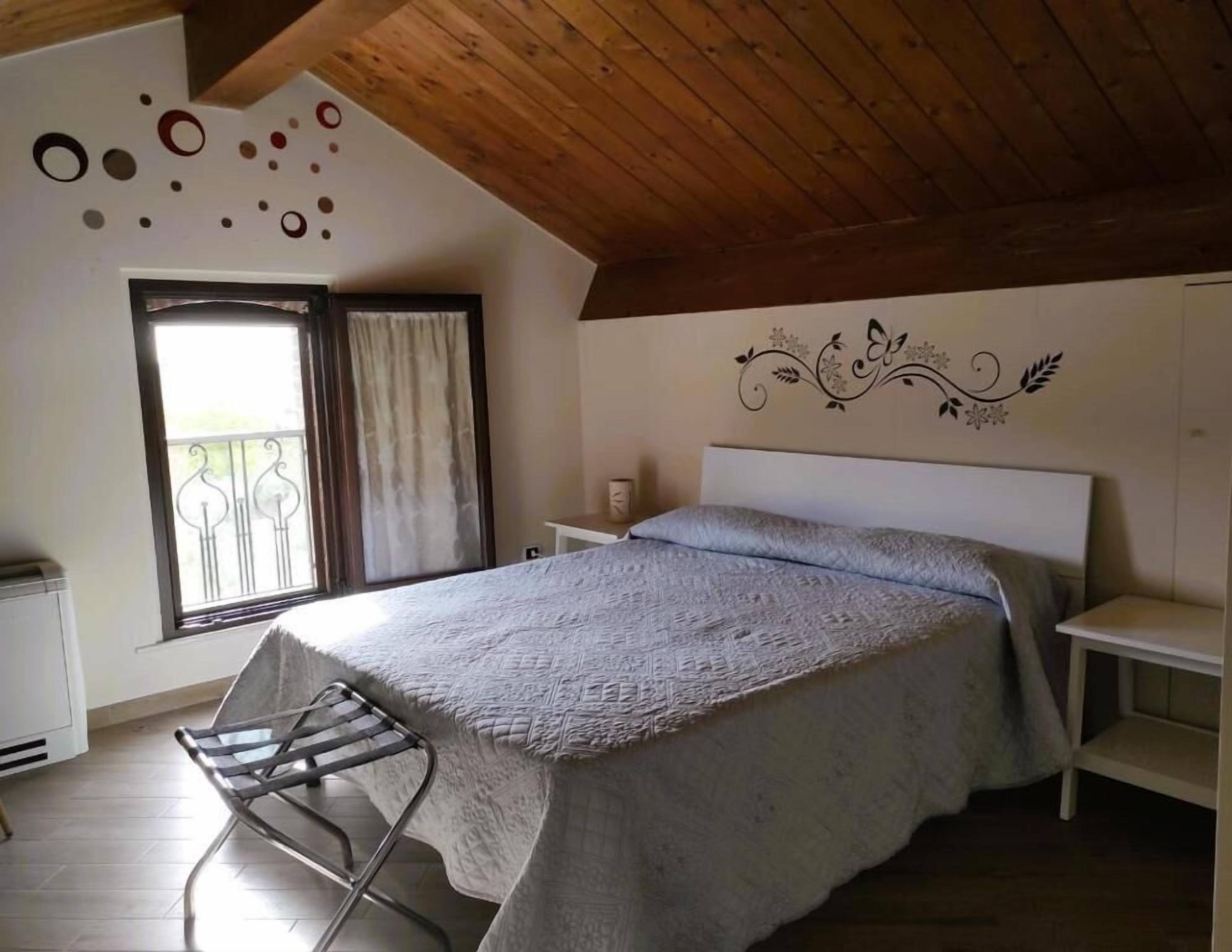 A bedroom with a bed, a chair, a window with curtains, a decorative wall, and a wooden ceiling.