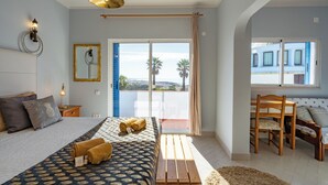 A bedroom with a bed, a wooden bench, a table with chairs, and a view of palm trees through the balcony doors. - Bem Parece by Umbral (Albufeira)