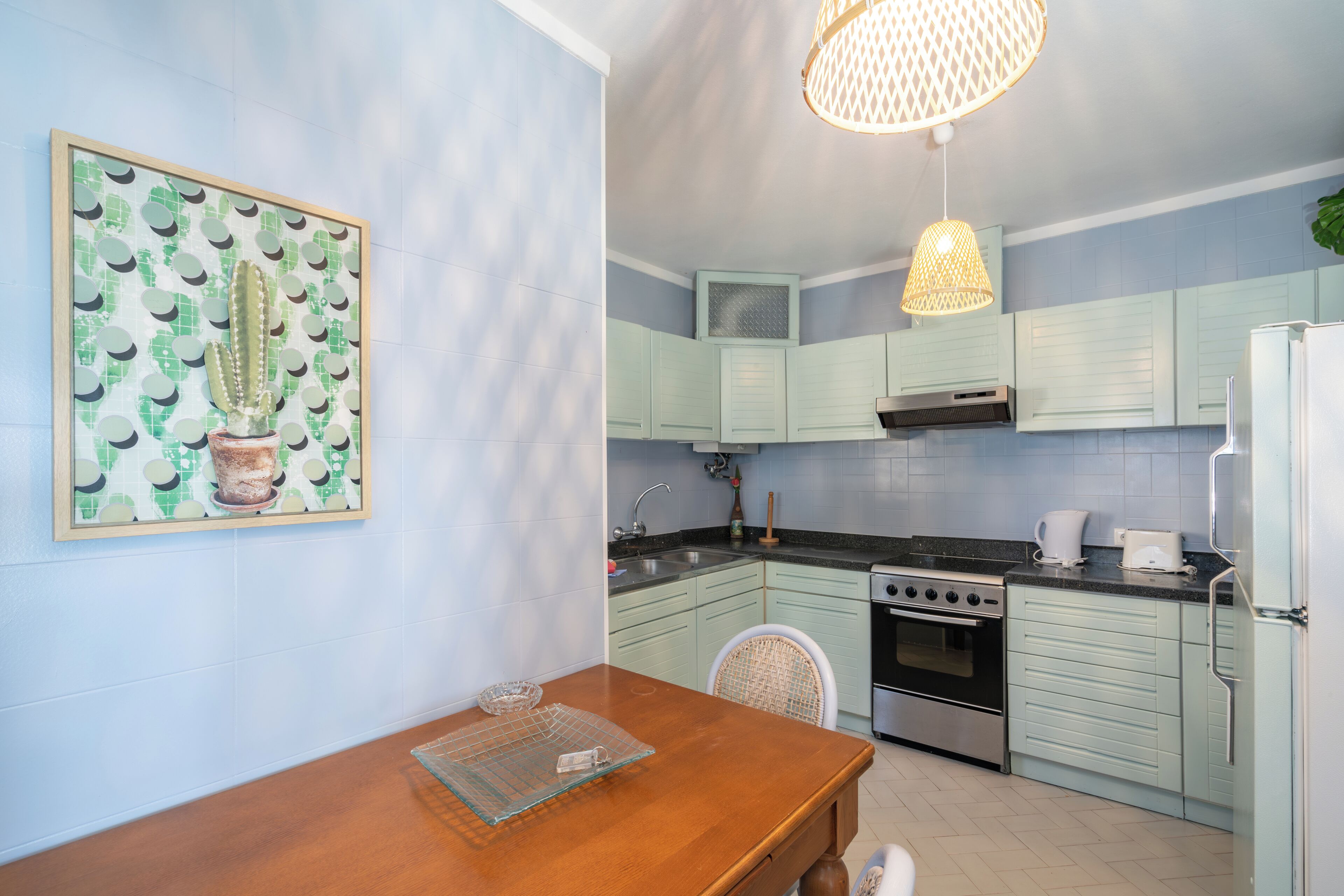 A kitchen with light blue cabinets, a wooden dining table, a hanging light fixture, and a framed cactus artwork on the wall.