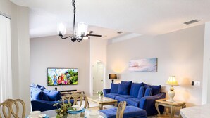 A modern living room with a dining area, featuring a TV, blue sofas, a chandelier, and a painting on the wall. - SD2216MC – Golf-side 5-Bed Villa with Private Pool (Haines City)