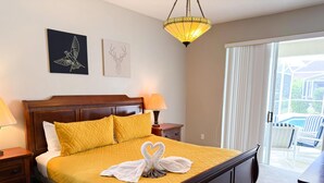 A neatly made bed with a yellow quilt, two pillows, and a swan-shaped towel. There are two wall-mounted artworks, a wooden nightstand with a lamp, and a view of an outdoor area through a glass door. - SD2216MC – Golf-side 5-Bed Villa with Private Pool (Haines City)