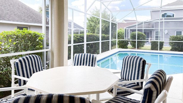 A sunlit patio with a pool, white-framed glass enclosure, and striped outdoor furniture. - SD2216MC – Golf-side 5-Bed Villa with Private Pool (Haines City)