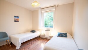 Una habitación pequeña y bien iluminada con dos camas individuales, una silla, un ventanal con vistas a zonas verdes y una lámpara en la pared.