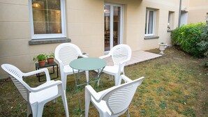 Un patio con sillas de plástico blancas y una mesa redonda pequeña, rodeado de un jardín con seto y planta en maceta.