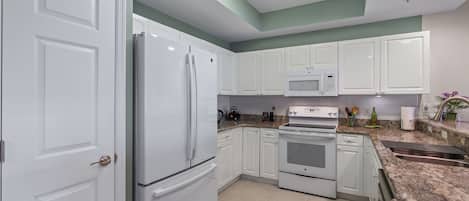 Una cocina con armarios blancos, refrigerador, horno, microondas y encimera de granito.