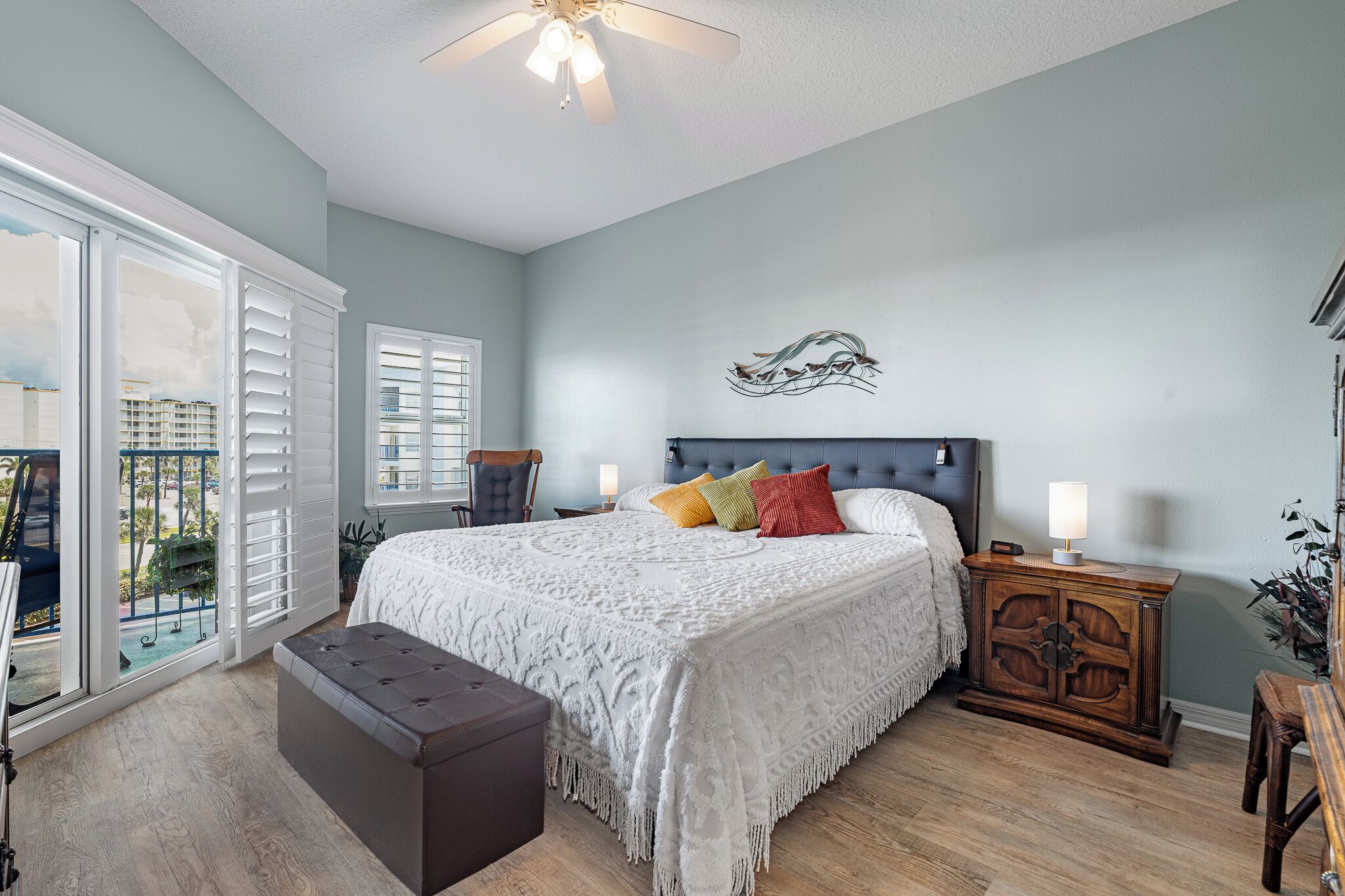 A bedroom with a large bed, a nightstand, a bench, and a view of the outdoors through a sliding door.