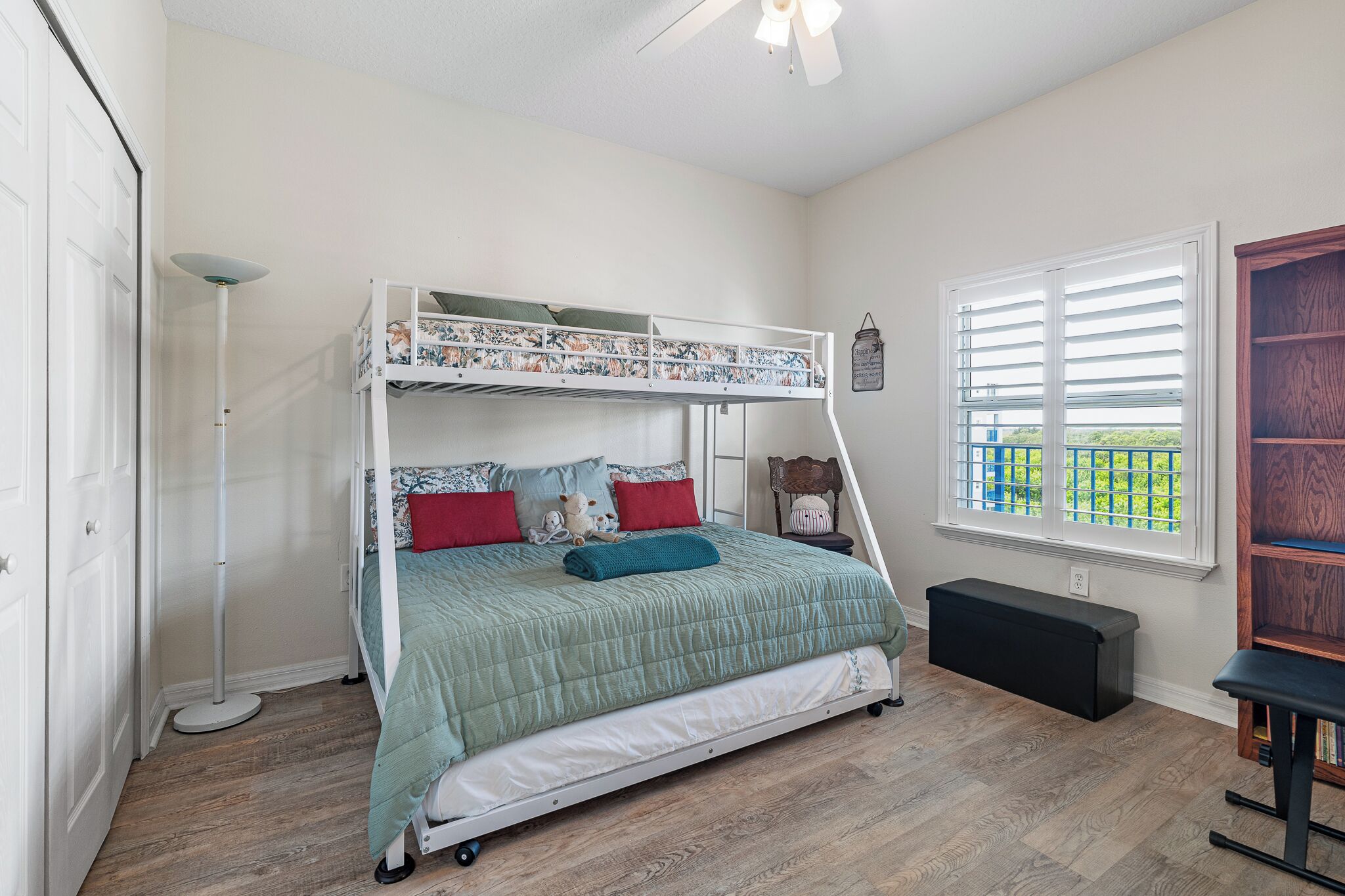 A bunk bed with a green quilt, red pillows, and a small bedside table with a teddy bear. There is a window with blinds, a floor lamp, and a black bench.