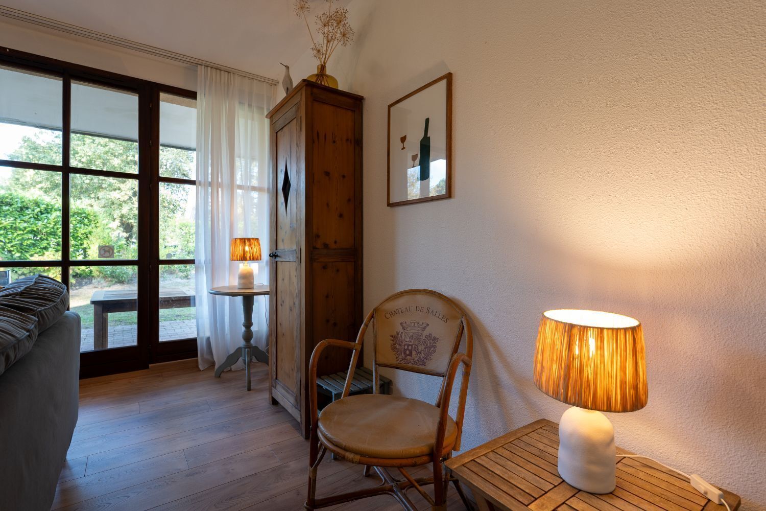 Una habitación con una silla de madera, una mesa con una lámpara, un armario de madera y una ventana con cortinas.