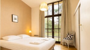 Una cama bien hecha con sábanas y toallas blancas, una lámpara de noche, una silla con cojín estampado y un ventanal con cortinas.