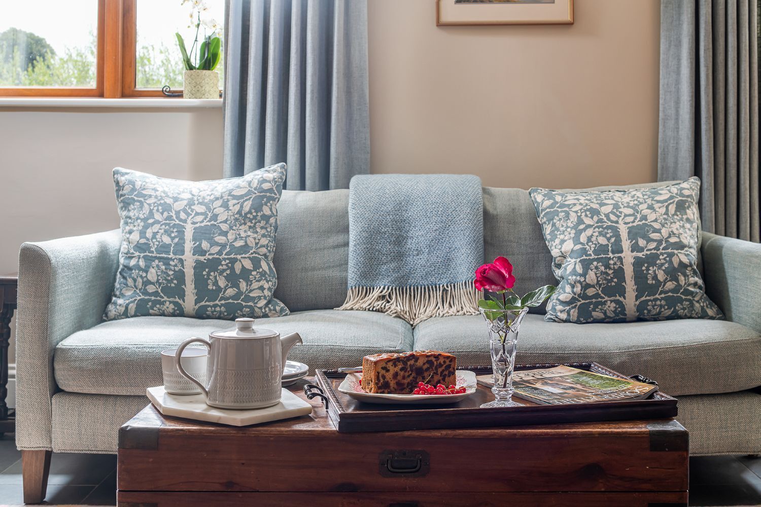 A living room with a sofa, pillows, a coffee table, a vase with a flower, and a tray with food and a book.