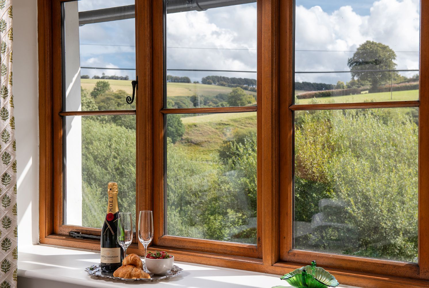 Una ventana con vistas a un paisaje verde, una botella de vino, dos copas y un cuenco con fruta sobre una bandeja.