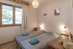 A room with two beds, a wooden nightstand, a small table, and a window with curtains. - Apartment in Samoëns – Sleeps 6, Near Slopes & Village (Samoëns)