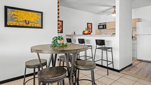A modern kitchen with white cabinets, a microwave, and a refrigerator. There is a dining area with a round table and four stools. A framed painting of a tree is on the wall. - Terrace Allows Pets Free Parking Otium (Fajardo)