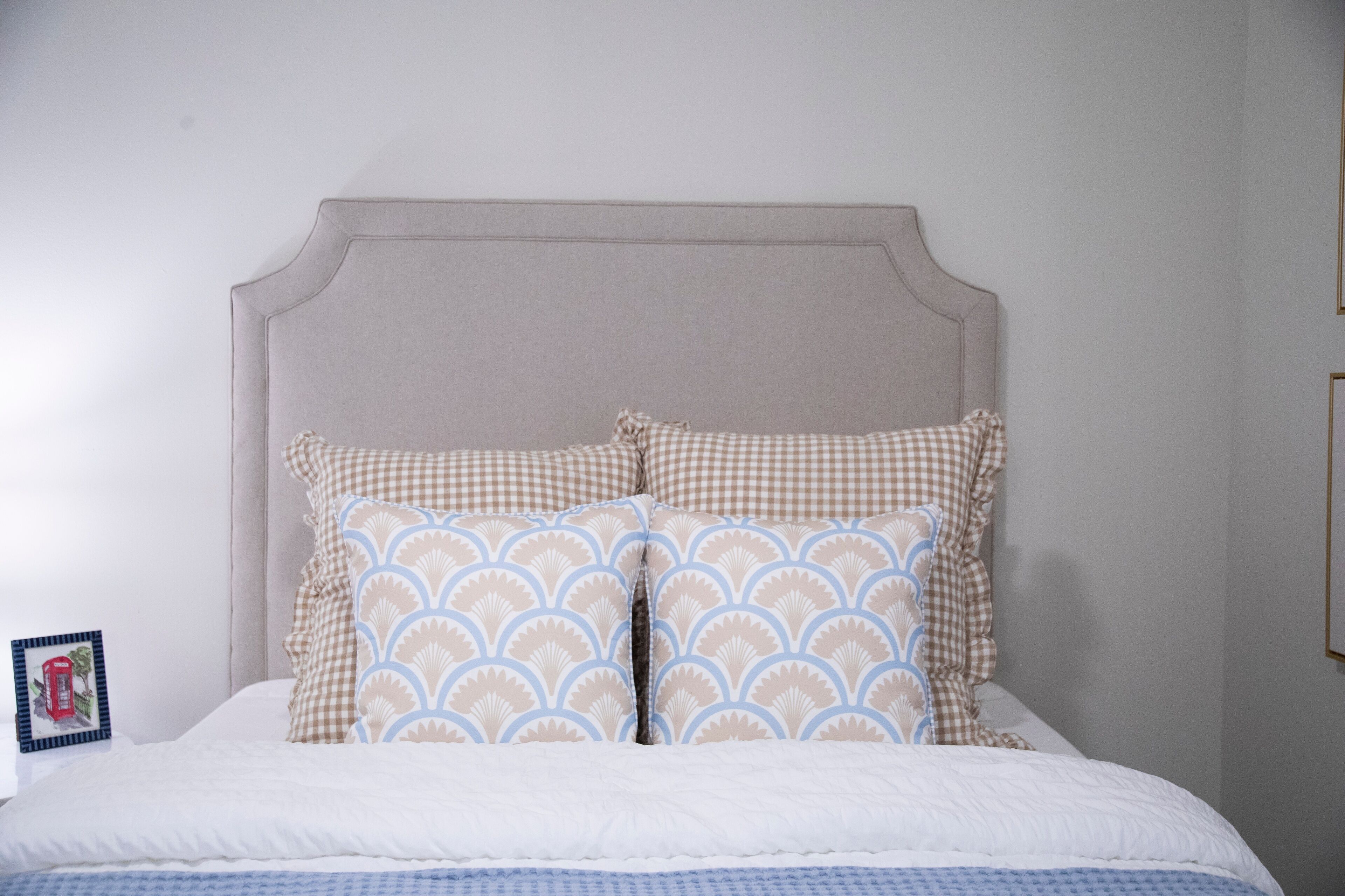 A bed with a checkered pillow and a patterned pillow, a framed picture on the nightstand, and a plain wall.